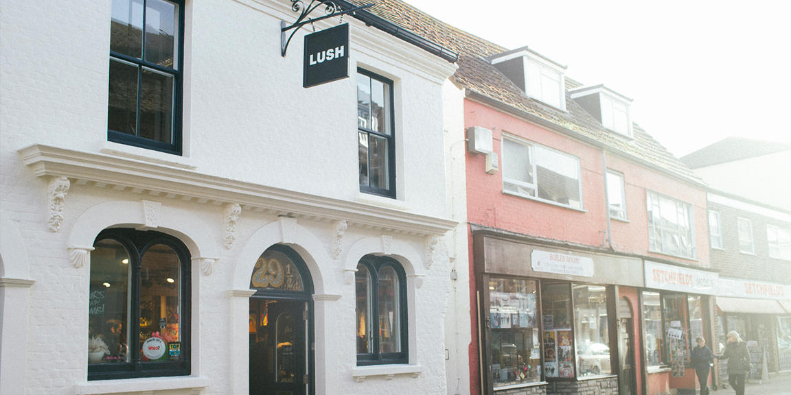 Exterior view of shop. White painted shopfront. Entrance in centre with with traditional hanging shop sign above. Products displayed in windows either side of entrance.