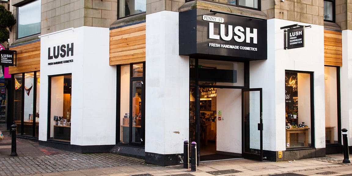 Lancaster shop front. Exterior view of shop. White framed shop. Large door way on right with illuminated Lush sign above. Products displayed in windows along side of shop.
