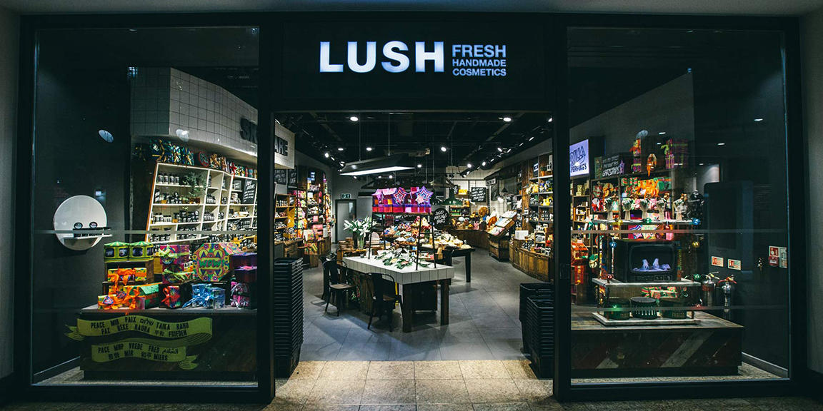 Meadowhall shop front. Exterior view of shop. Black framed shop with glass frontage. Large door way in centre with illuminated Lush sign above. Products displayed in windows either side of entrance.
