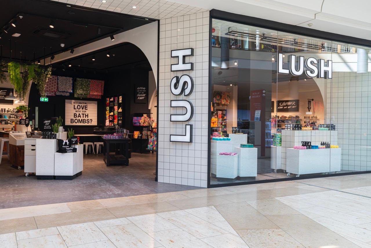 Exterior view of shop. Central tiled column with illuminated Lush sign. Large doorway to left hand side with full view of shop interior. Large glass window to the right of the entrance with several product displays.