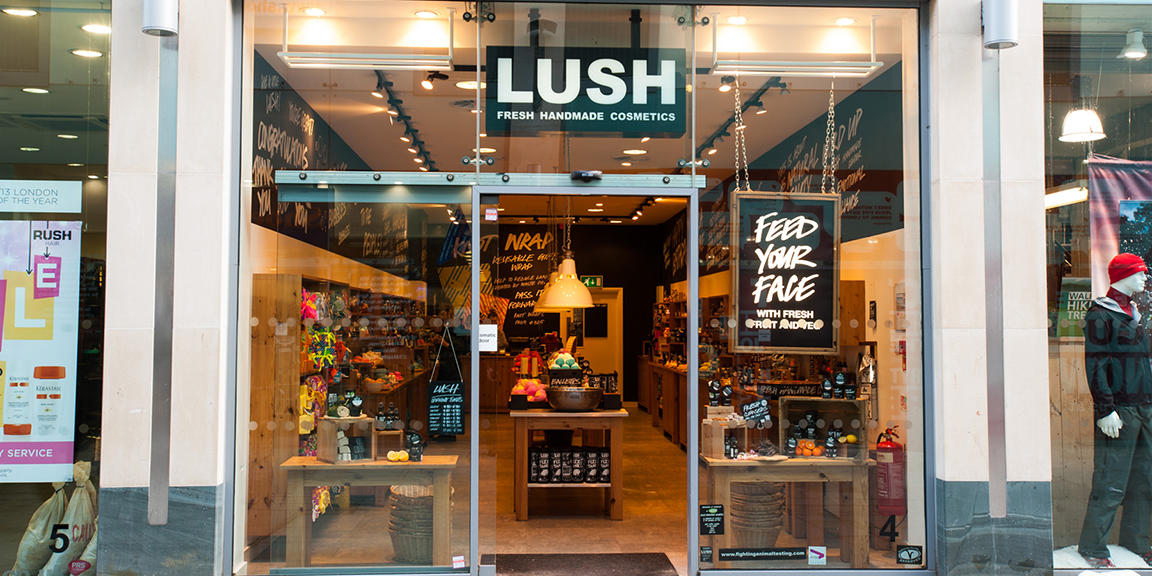 Exterior view of shop. Glass fronted shop. Door way in centre with illuminated Lush sign above. Products displayed in windows either side of entrance.