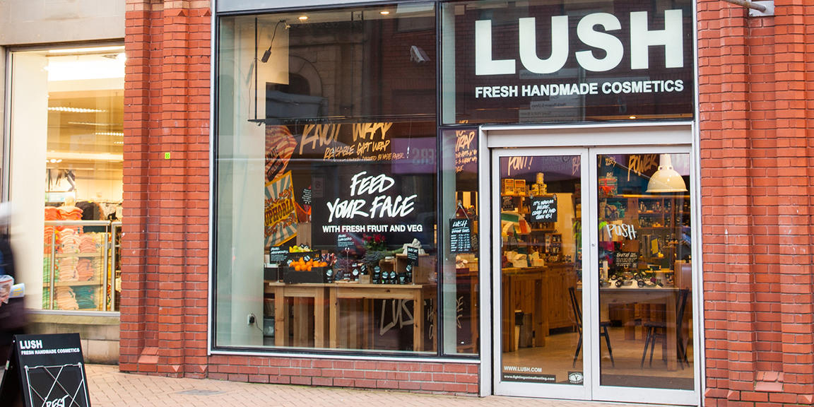 Preston shop front. Exterior view of shop. Glass fronted shop. Large entrance on right with illuminated Lush sign above. Products displayed in window to left side of entrance.