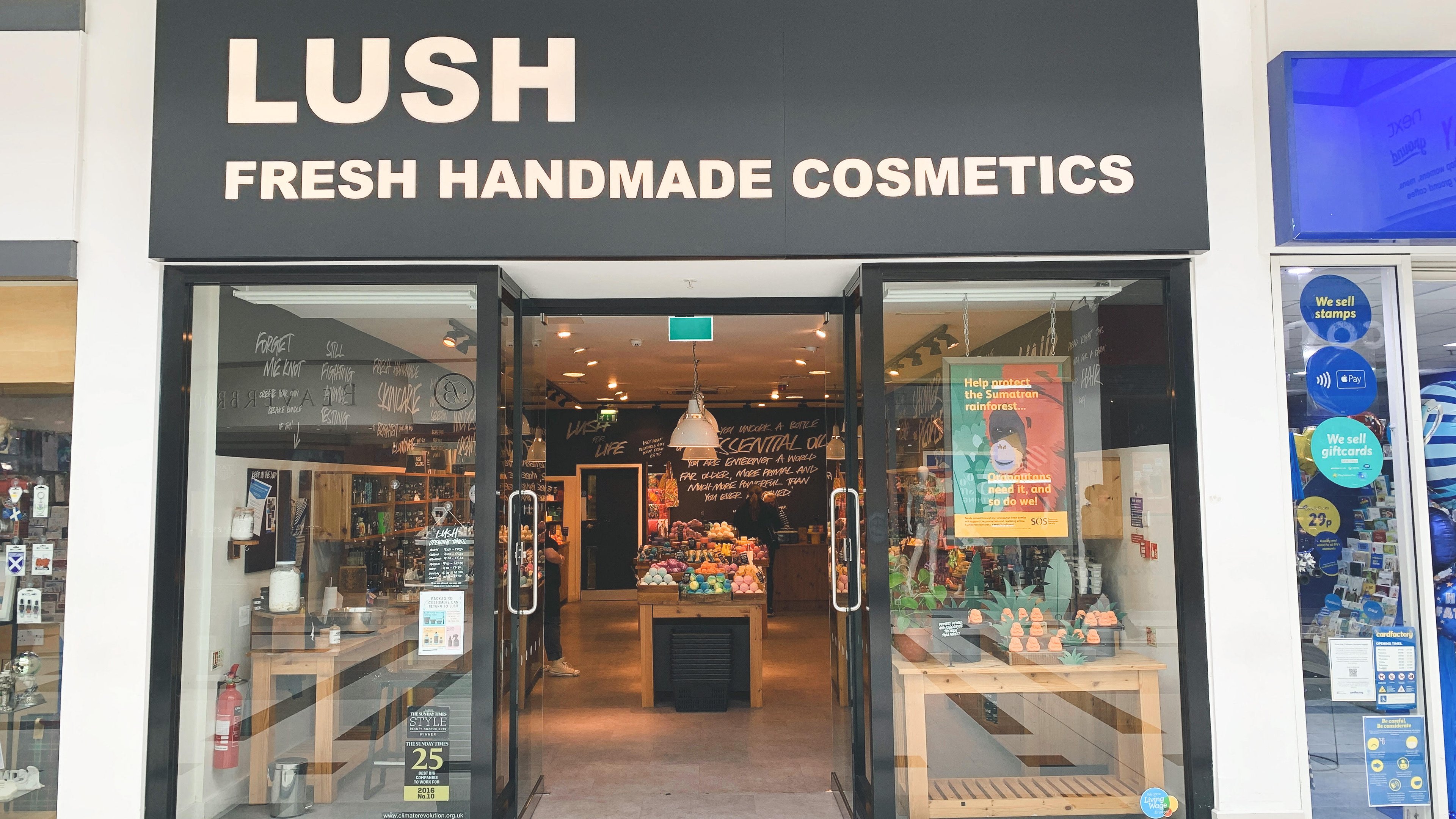 Exterior view of shop. Black framed shop with glass frontage. Entrance in centre with illuminated Lush sign above. Products displayed in window to the right side of entrance underneath an illuminated lightbox.