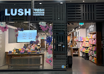 Exterior view of shop. Black framed shop with glass frontage. Large door way on right. Products displayed in windows on left side of entrance with illuminated Lush sign above.