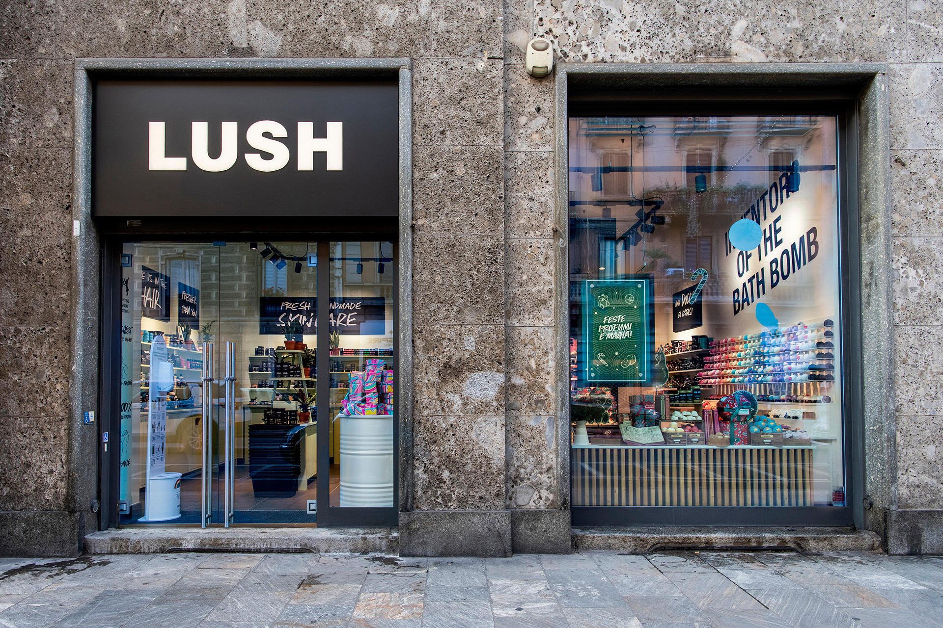 Exterior view of shop. Grey stone framed shop. Entrance on the left with illuminated Lush sign above. Products displayed in window on right side of entrance.