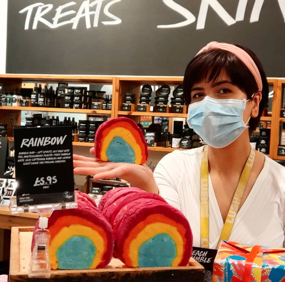 Member of staff wearing a facemask in middleground. Rainbow coloured unpackaged bath products in foreground.