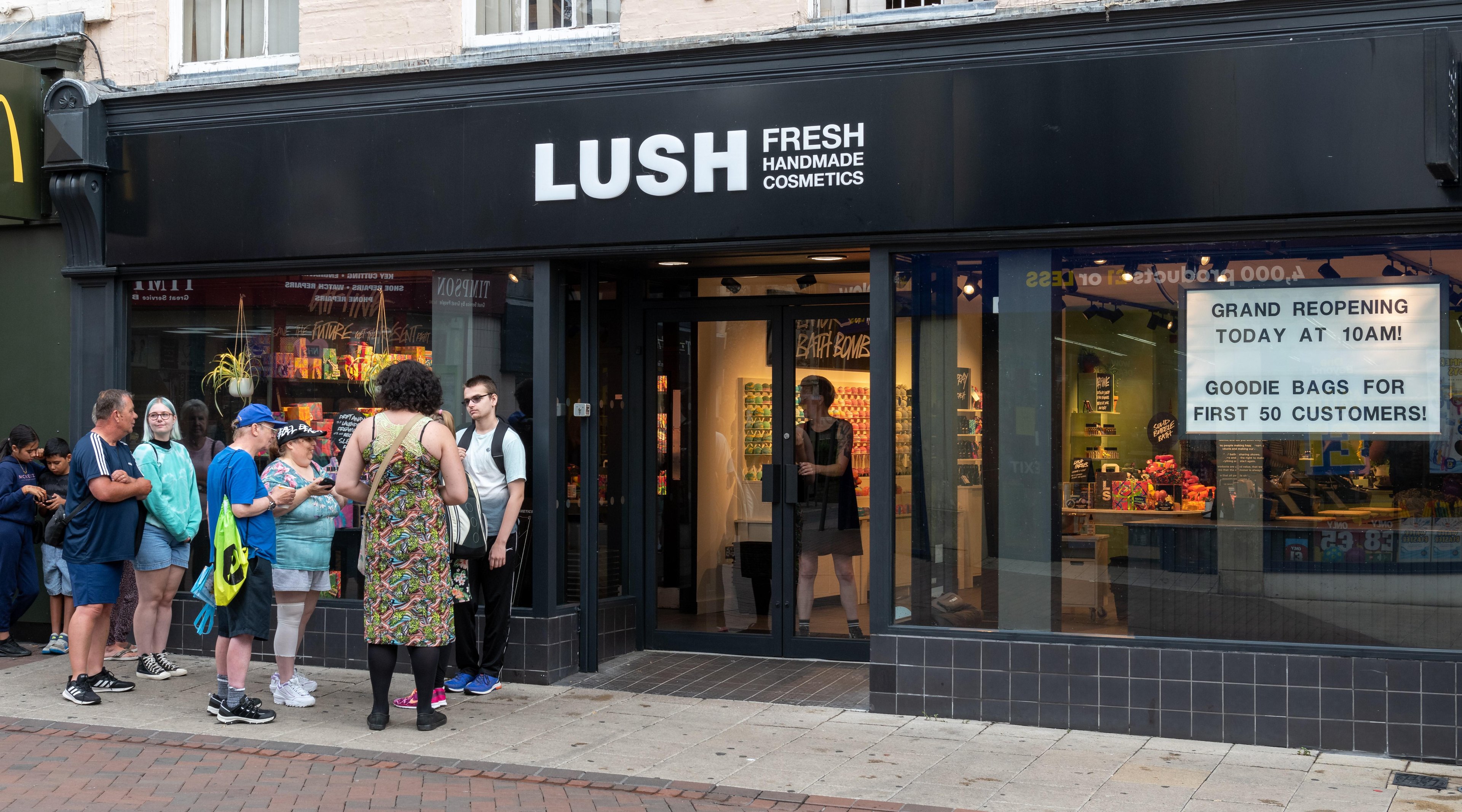 Exterior view of shop. Black framed shop with glass frontage. Large door way in centre with illuminated Lush sign above. Customers queueing on left hand side of shop to enter.