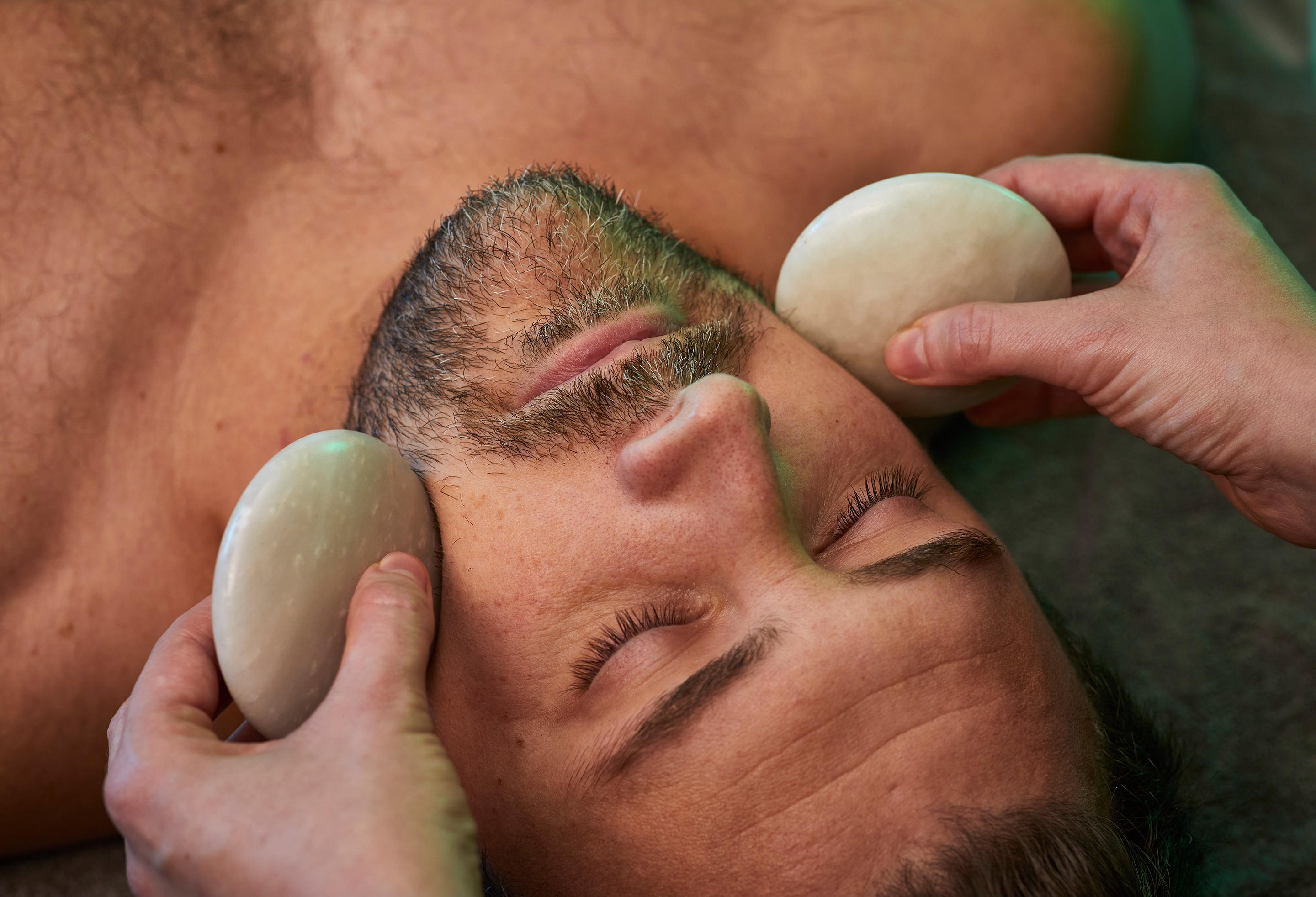 Person with facial hair having two stones held to the sides of the face by a therapist.