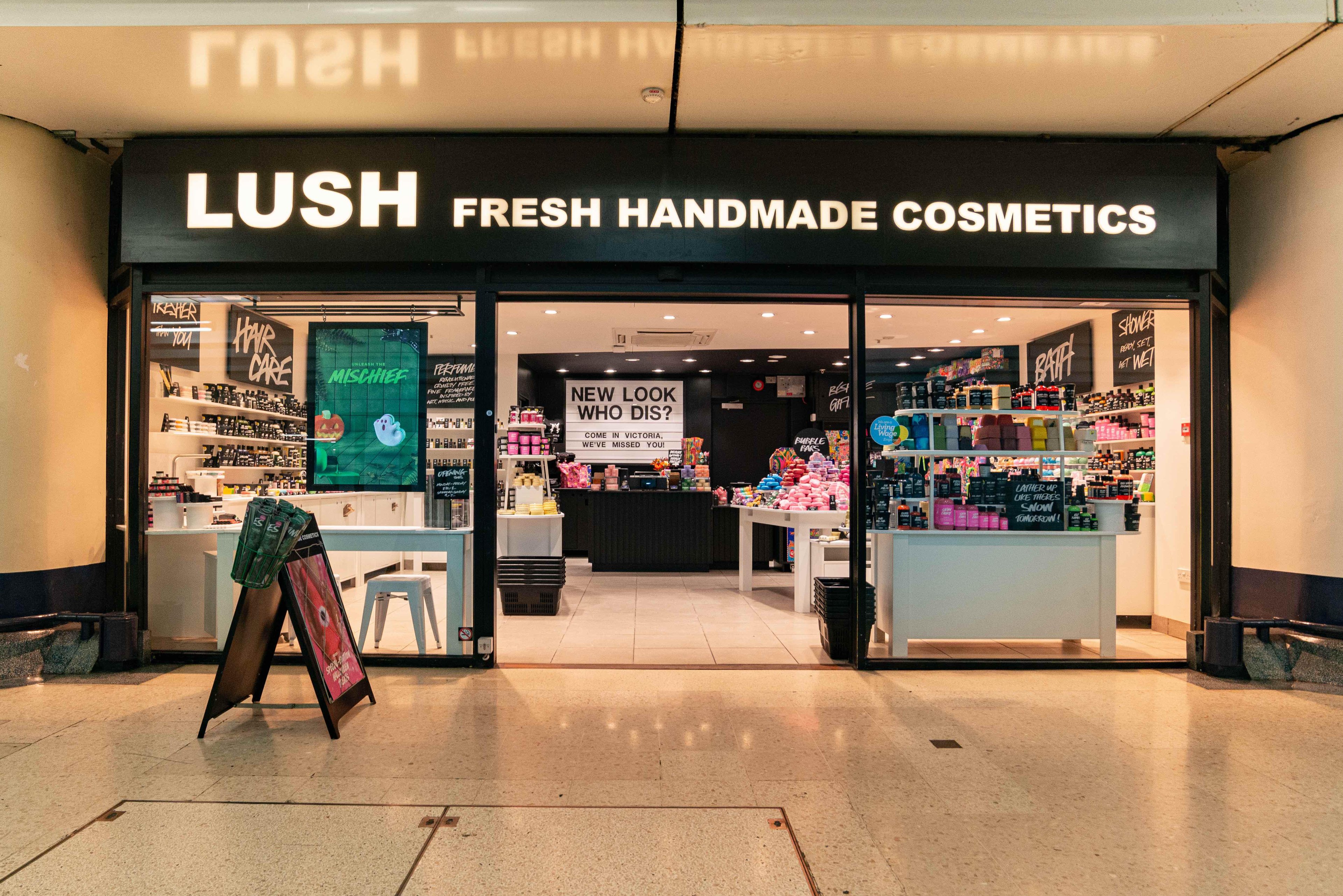 Exterior view of shop. Black framed shop with glass frontage. Large entrance in centre with illuminated Lush sign above. Products displayed in windows either side of entrance.