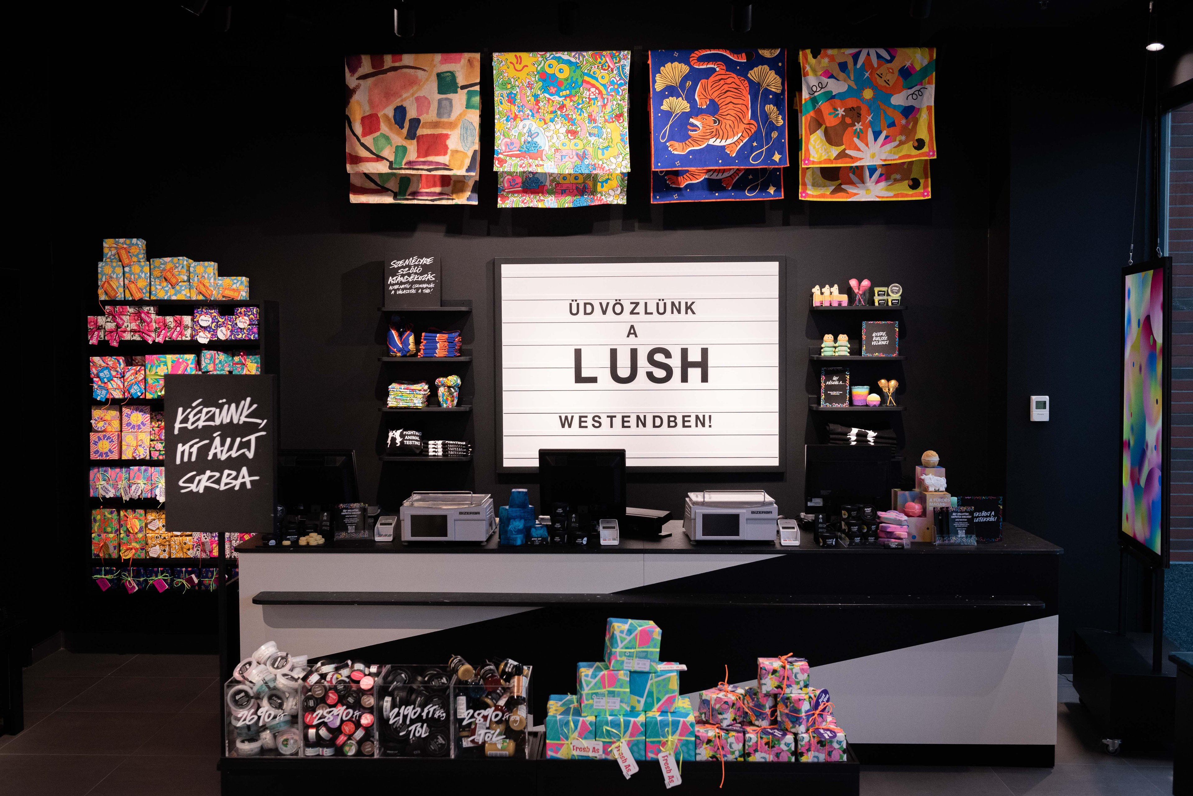 Interior of the shop looking at the till point. Large cinema style display board behind the counter. Colourful gift display to the left of the counter. Knotwrap scarves hanging from the ceiling above the counter.