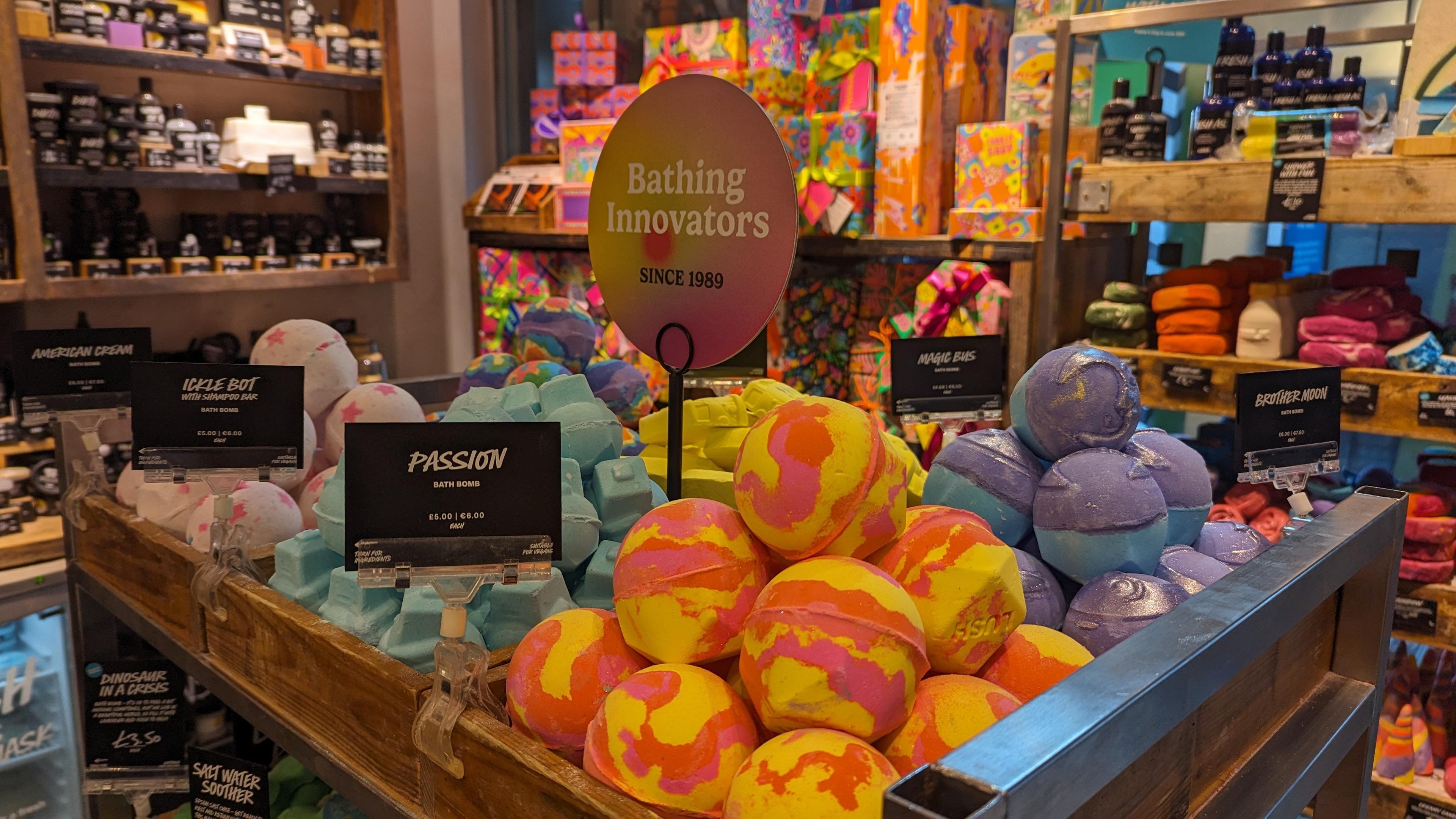 Interior view. large display of unpackaged colourful bath products in wooden pens. Sign above reads Bathing Innovators.