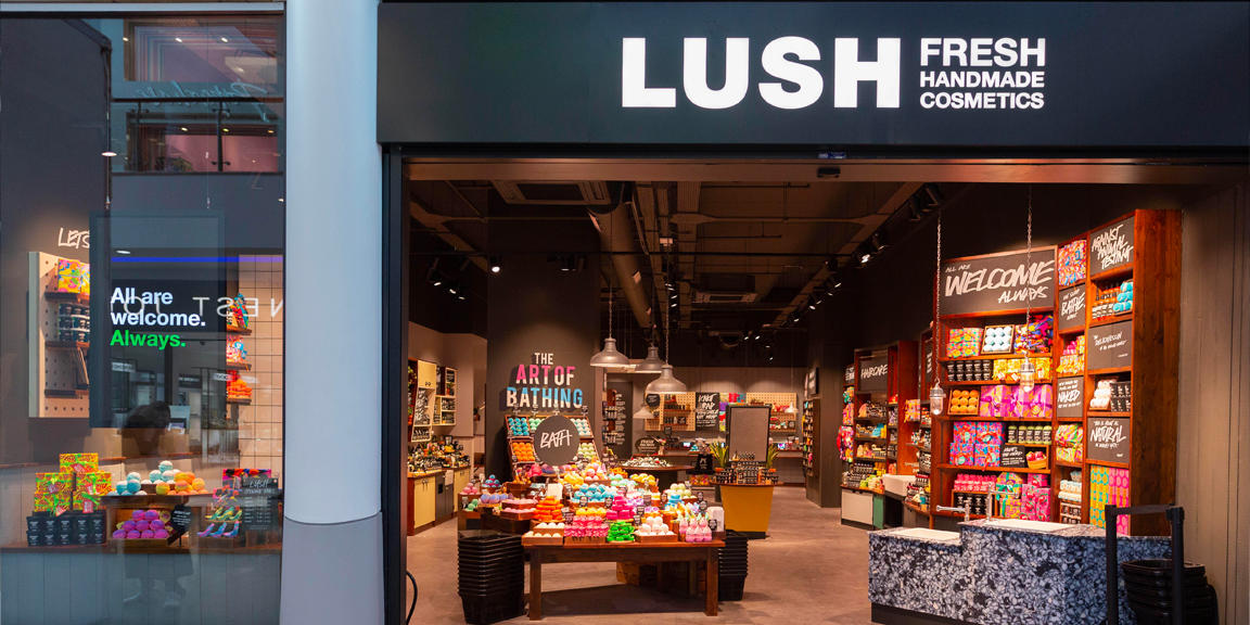 Exterior view of shop. Black framed shop with glass frontage. Very large entrance to the right with illuminated Lush sign above. Products displayed in window to left side of entrance.
