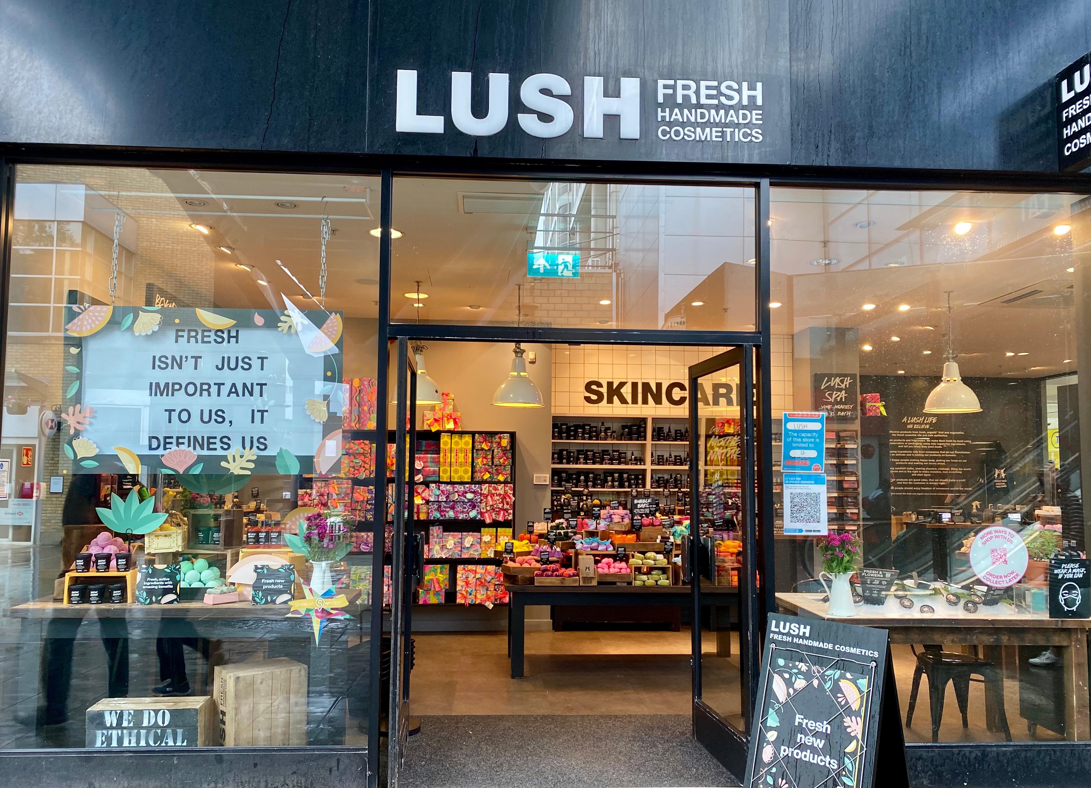 Exterior view of shop. Black framed shop with glass frontage. Large entrance in centre with illuminated Lush sign above. Products displayed in windows either side of entrance.