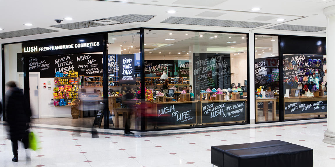 Exterior view of shop. Black framed shop with glass frontage. Large door way on left with illuminated Lush sign above. Products displayed in windows along side full shop frontage.