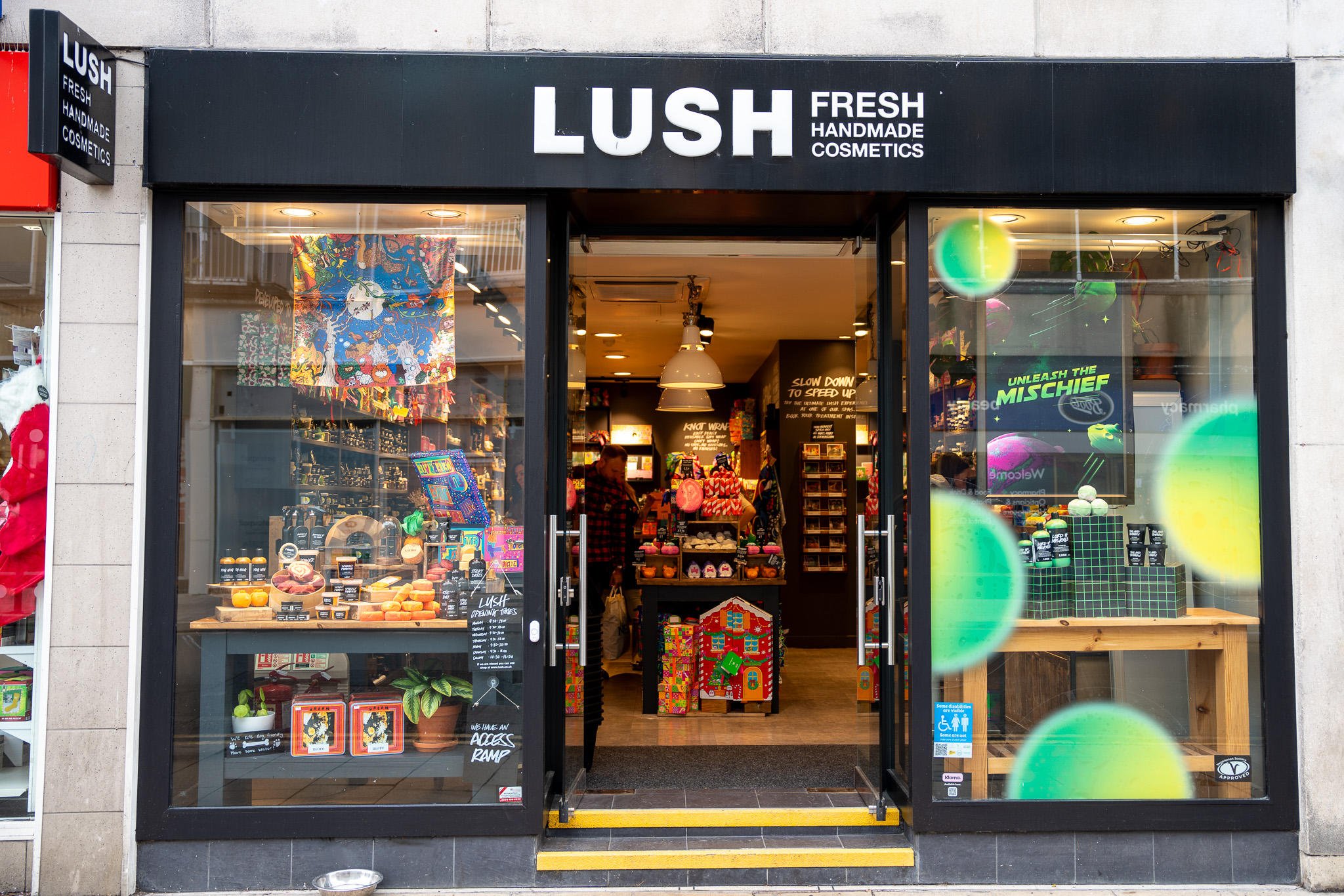 Exterior view of shop. Black framed shop with glass frontage. Door way in centre with illuminated Lush sign above. 3 steps up into the shop. Products displayed in windows either side of entrance.
