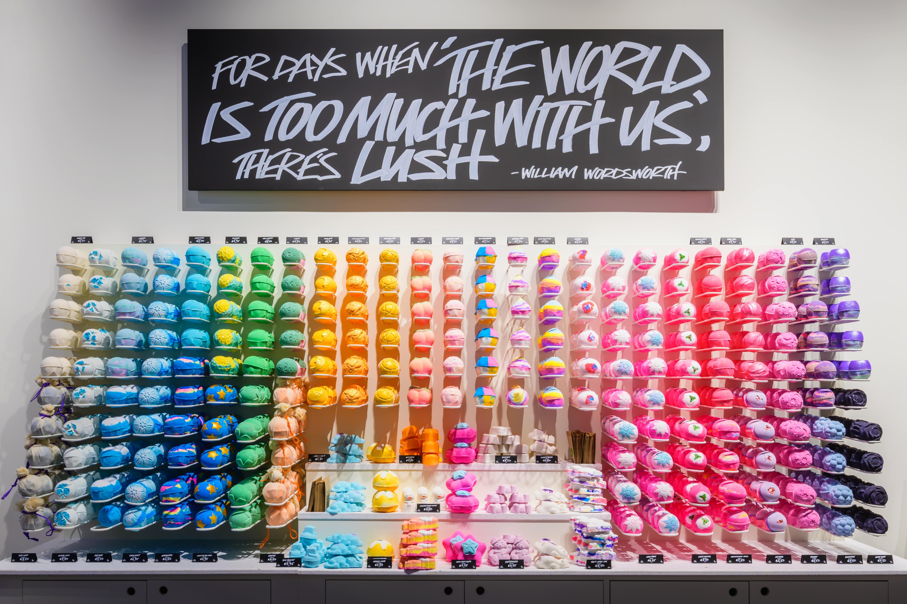 Very colourful display of unpackaged bath products. Products are displayed in columns of colours creating a rainbow like effect from left to right. Sign above reads "For days when the world is too much with us" there's Lush.