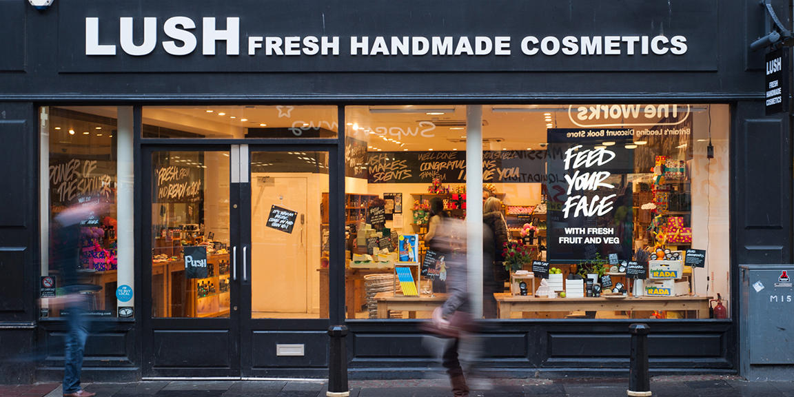 Lush Inverness shop front. Exterior view of shop. Black framed shop with glass frontage. Large door way to left of view with illuminated Lush sign above. Products displayed in windows to right of entrance.