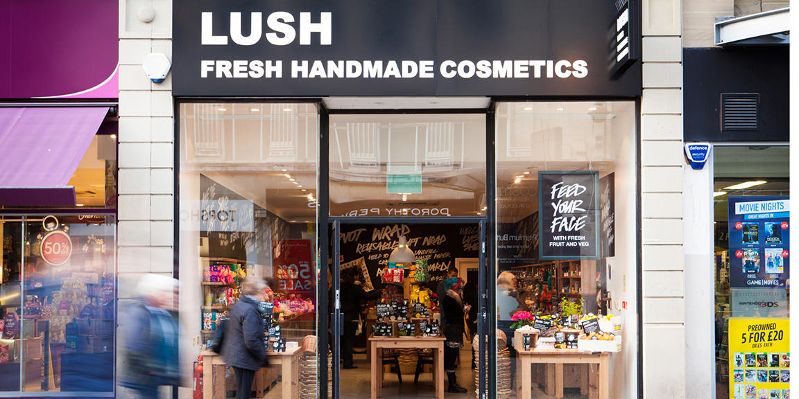 Sheffield shop front. Exterior view of shop. Black framed shop with glass frontage. Entrance in centre with illuminated Lush sign above. Products displayed in windows either side of entrance.