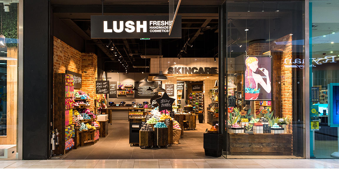 Derby shop front. Black framed shop with large doorway. Illuminated Lush sign above. Product display in window to the right of the entrance. Full interior of the shop can be seen.