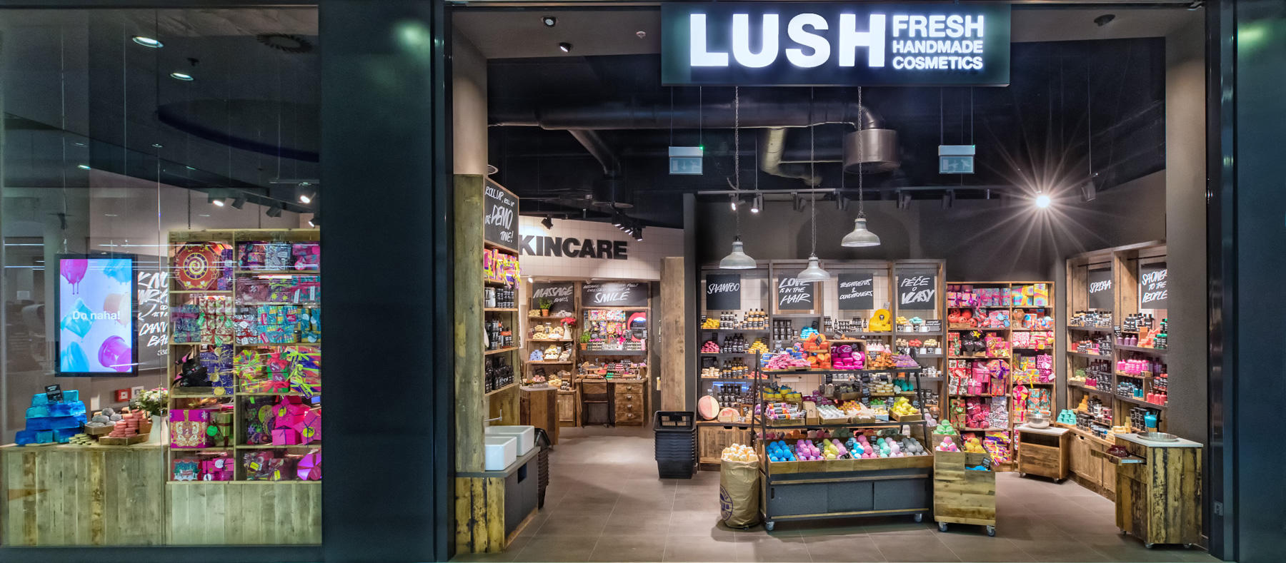 Exterior view of the shop. Large open doorway on the right with illuminated LUSH sign above. Full view inside the well lit shop. Large glass window on the left of the door with an illuminated lightbox display and products beneath.