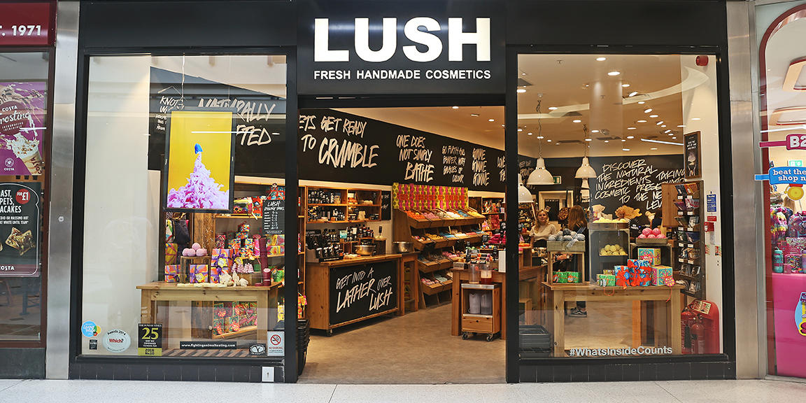 Watford shop front. Exterior view of shop. Black framed shop with glass frontage. Large entrance in centre with illuminated Lush sign above. Products displayed in windows either side of entrance.