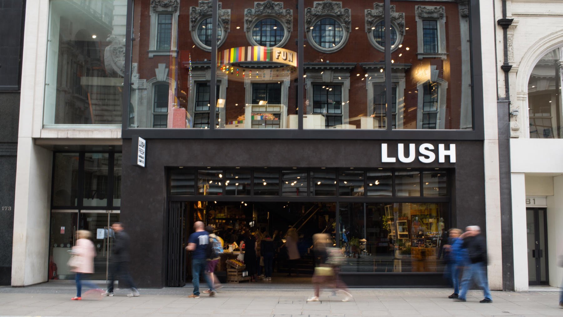 Lush Oxford Street shop front. Exterior view of shop. Black framed shop with glass frontage. Large entrance on left side with illuminated Lush sign above on the right. Products displayed in window to the right side of entrance.