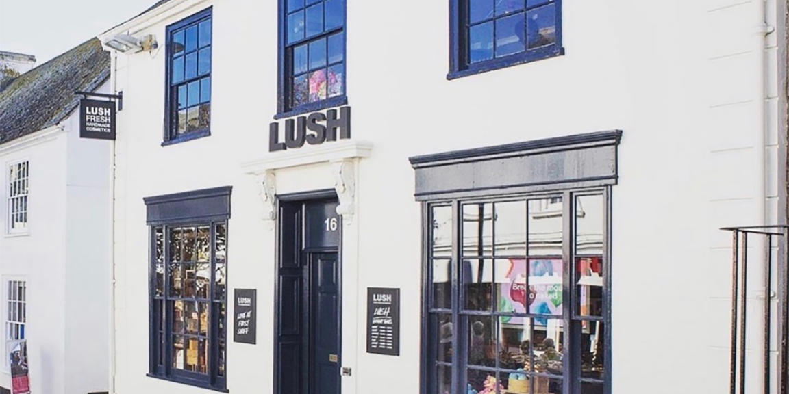 Truro shop front. Exterior view of shop. White traditional shopfront with black window frames and doorframe . Entrance in centre with Lush sign above. Step up into the shop. Products displayed in windows either side of entrance.