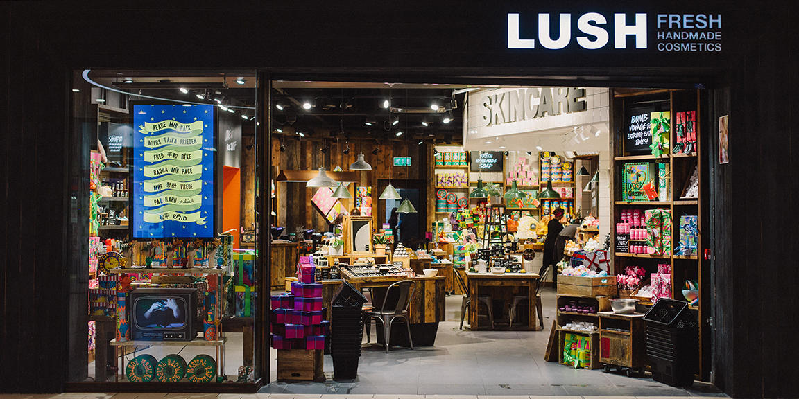Exterior view of shop. Black framed shop with glass frontage. Large door way on the right with illuminated Lush sign above. Products displayed in window on left of entrance.