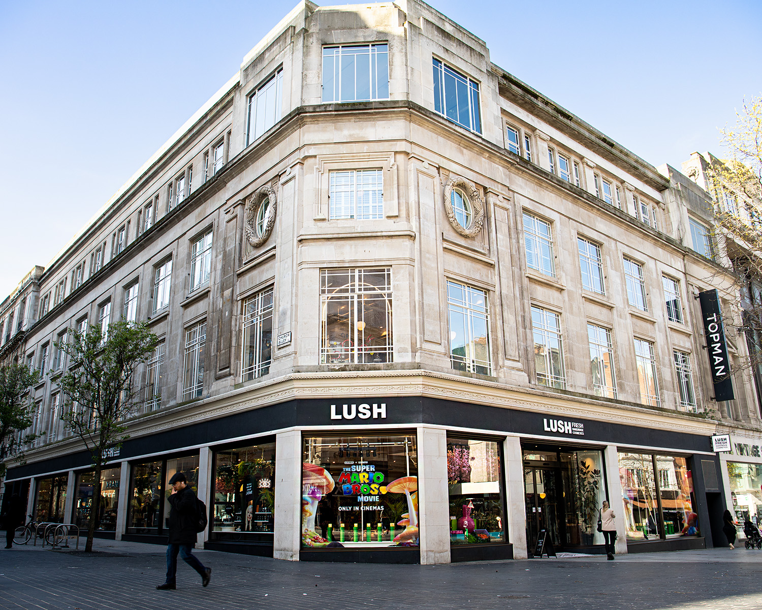 Exterior view of shop. White stone corner building consisting of 4 storeys. Large entrance to the right with illuminated Lush sign above. Products displayed in windows along both lengths of the shop.