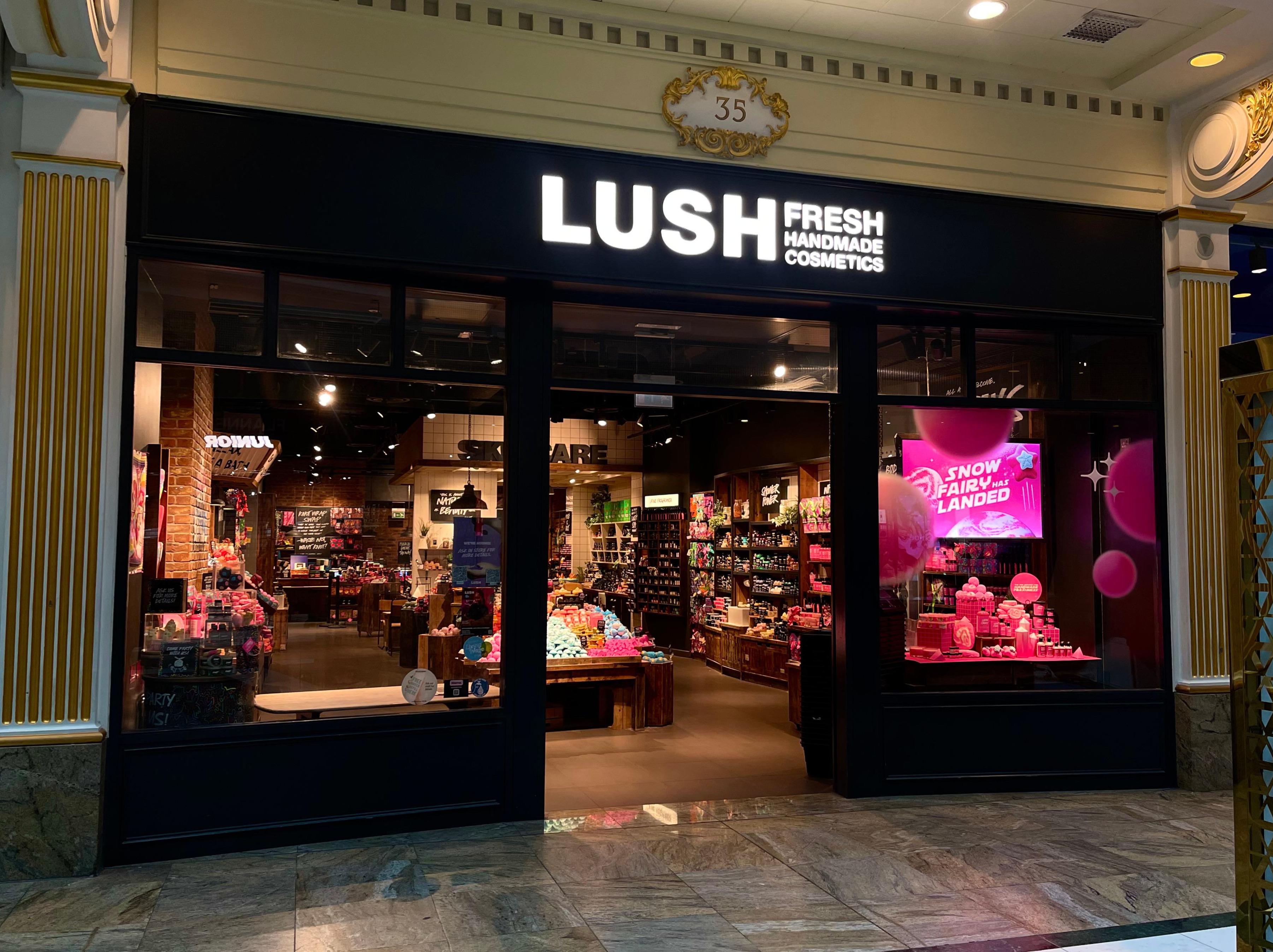 Exterior view of shop. Black framed shop with glass frontage. Large door way in centre with illuminated Lush sign above. Products displayed in windows either side of entrance.