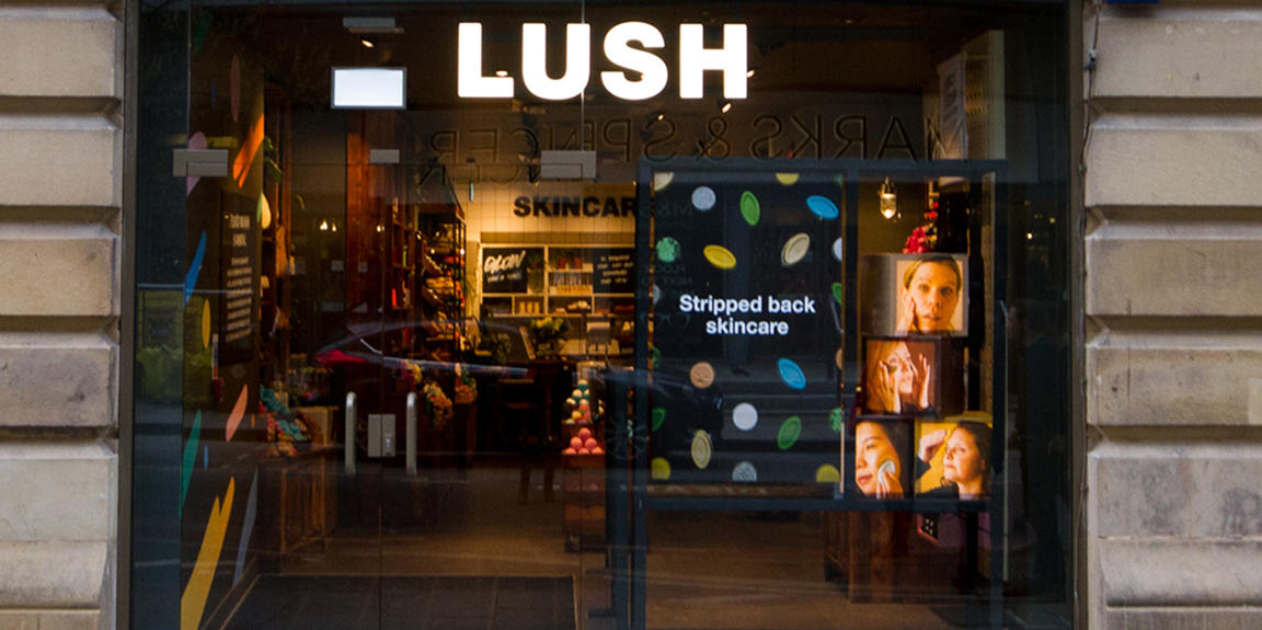 Manchester Market Street shop front. Exterior view of shop. Black framed shop with glass frontage. Door way on left with illuminated Lush sign above. Products displayed in window on right side of entrance.