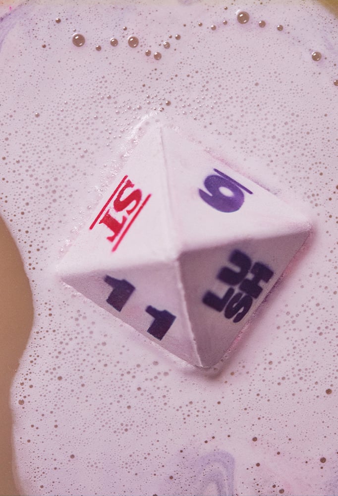 D8 bath bomb, in the shape of an 8 sided dice, floats upwards in water covered by white bubbles.