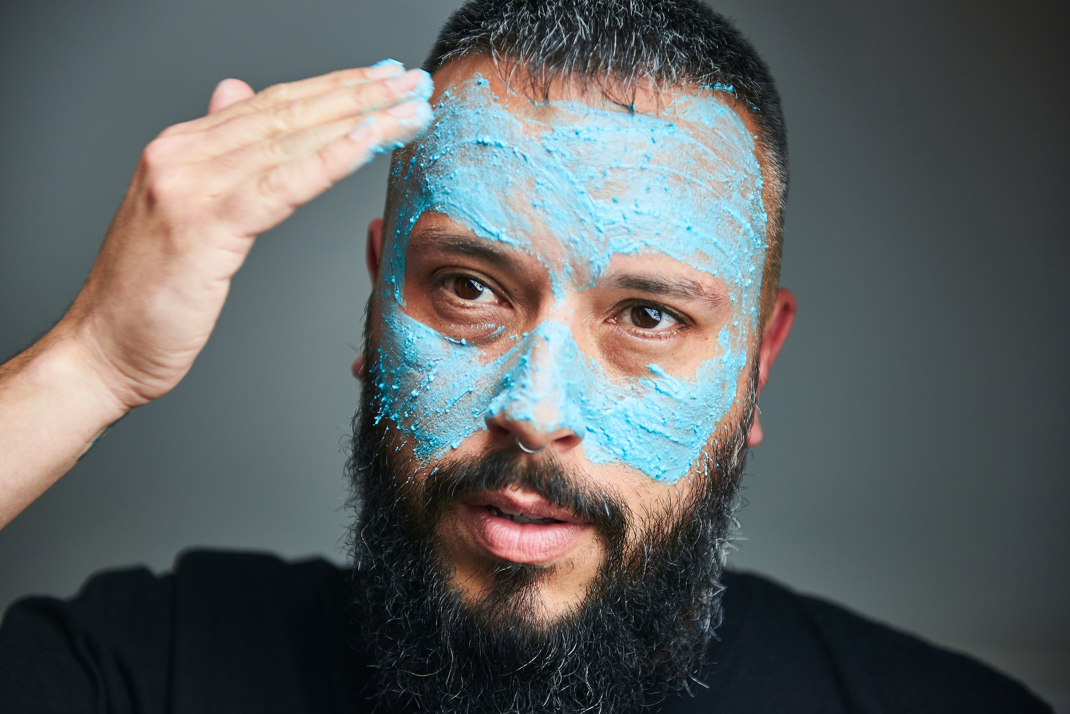 The bearded model gently applies the fresh, aqua-blue face mask to their face with their fingertips, on a grey background.