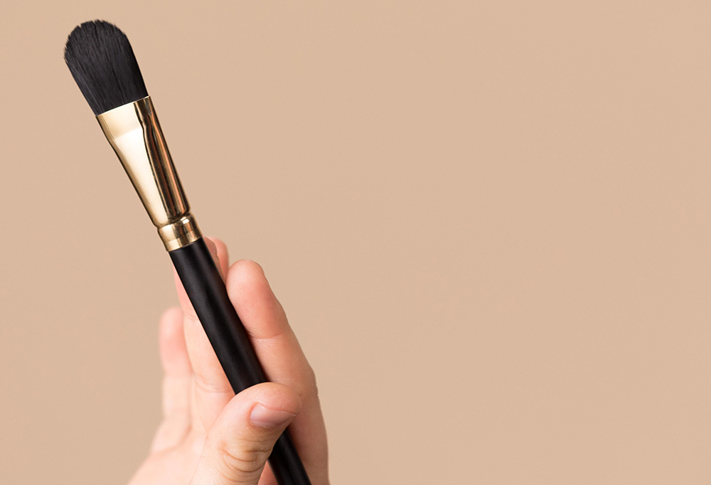 A hand holds a Fresh Faced Brush - a flat foundation/ face mask brush, with tapered brown bristles.