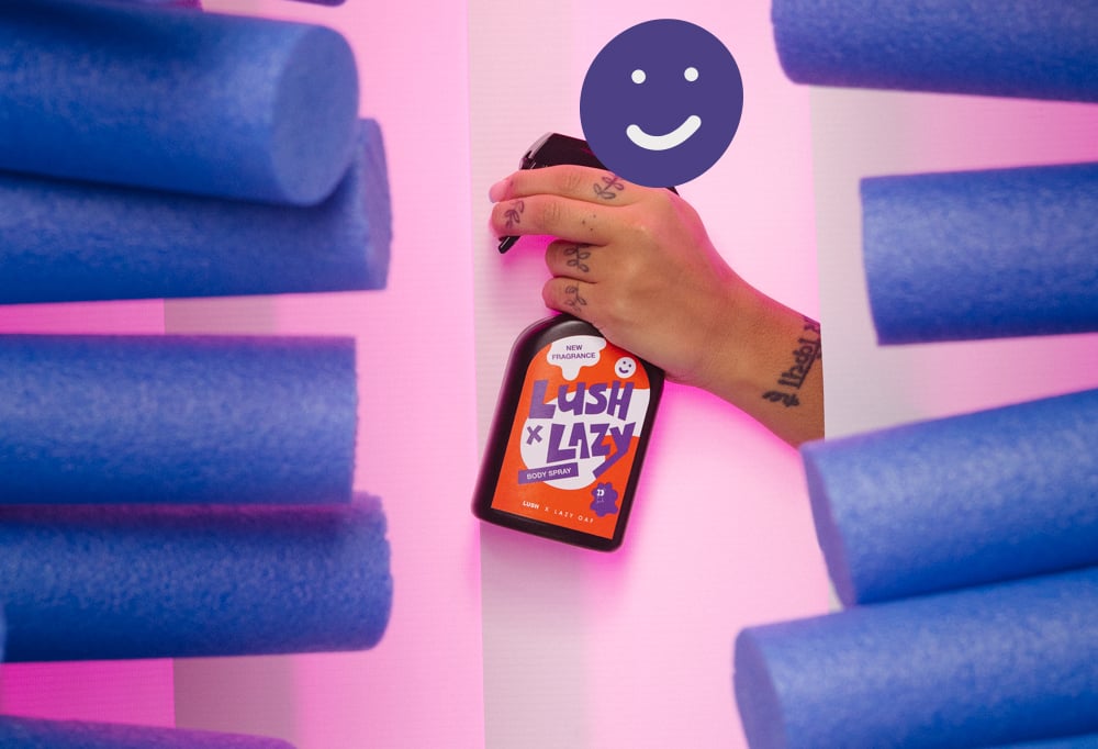 Lush x Lazy body spray is held the air, between blue foam pool floats on a vibrant pink background.