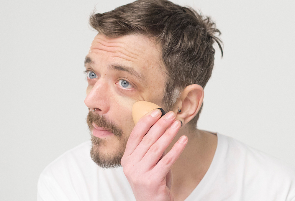 A bearded person leans foward as they apply a light, egg-shaped solid foundation Slap Stick to their under eye and cheek area.