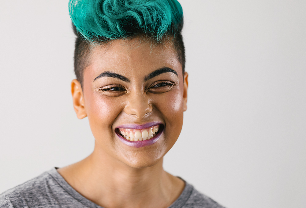 A person with short, turquoise blue hair grins cheesily, showing off dewy looking made up skin.