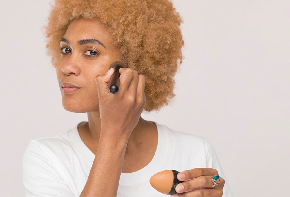A person with blonde afro hair uses a BFF make up brush to apply a medium dark-warm, egg-shaped solid foundation Slap Stick.