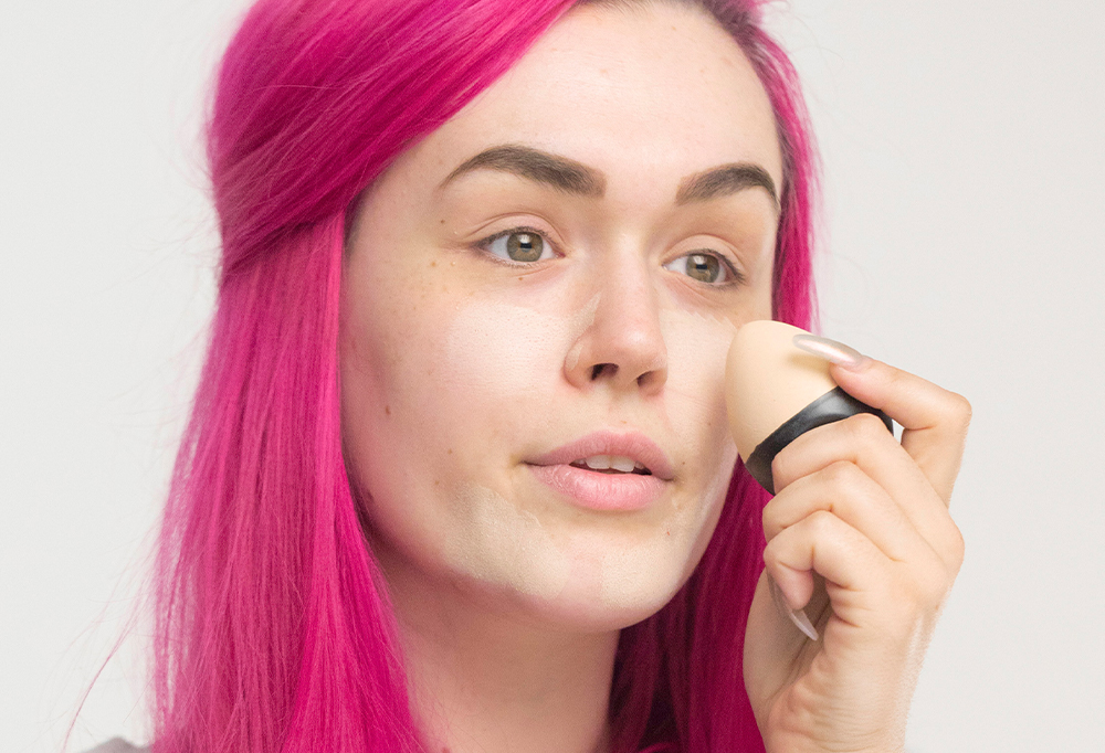 A person with vibrant pink hair applies a light, egg-shaped solid foundation Slap Stick all over their face.