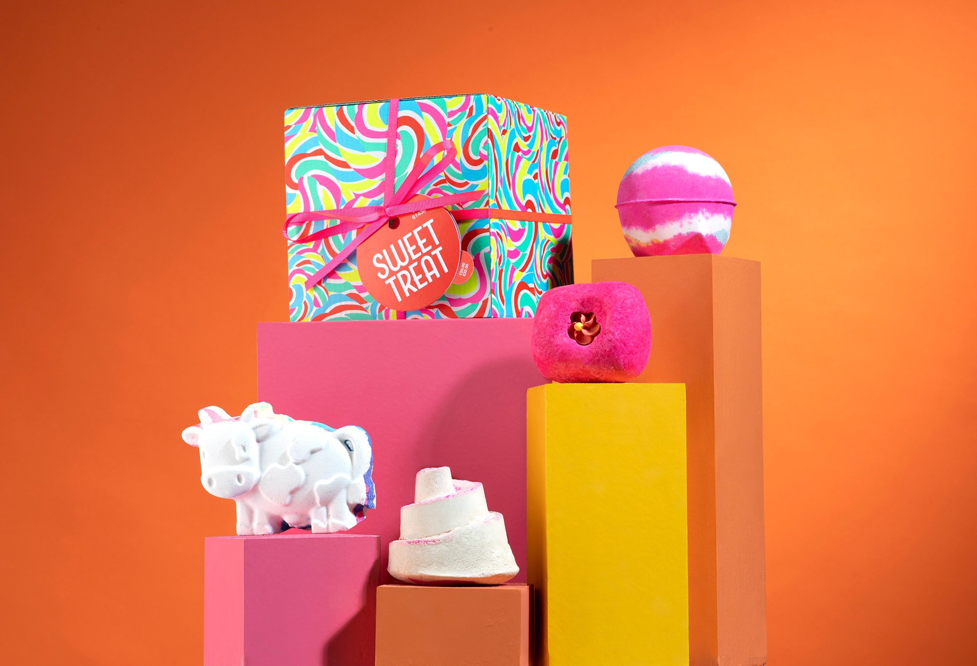 Sweet Treat gift, in front of an orange background, surrounded by its products on pink, orange and yellow plinths.