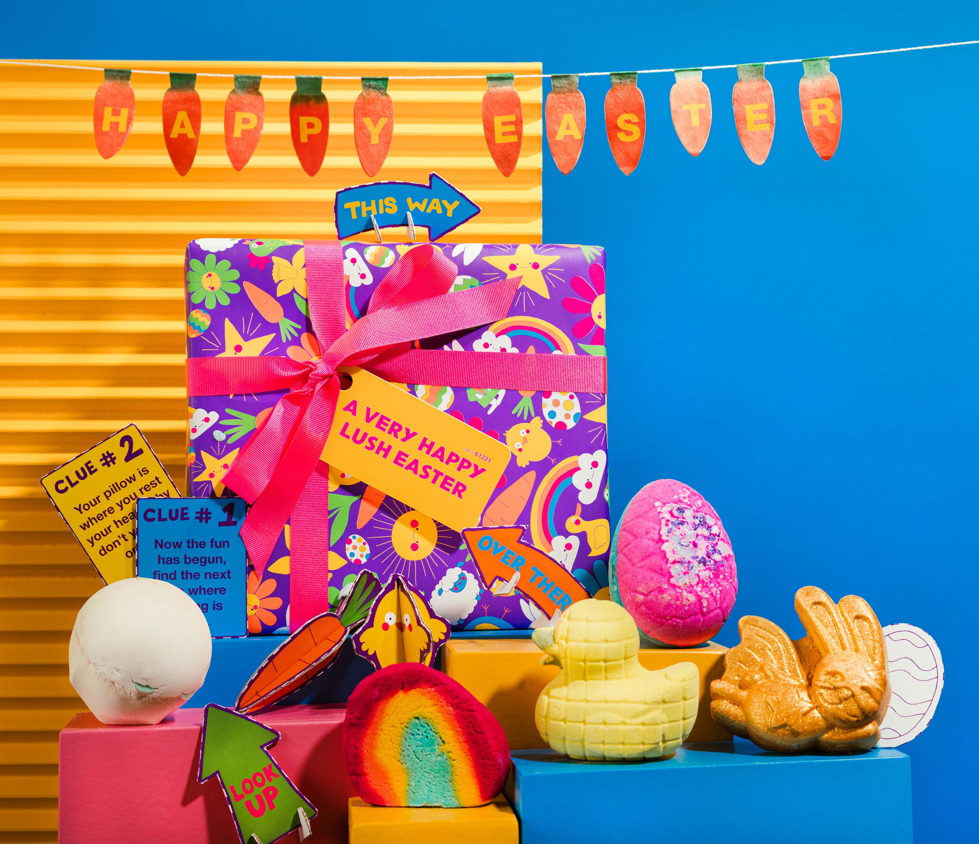 The gift in front of a blue and yellow background, surrounded by its products on blue, yellow and pink plinths.