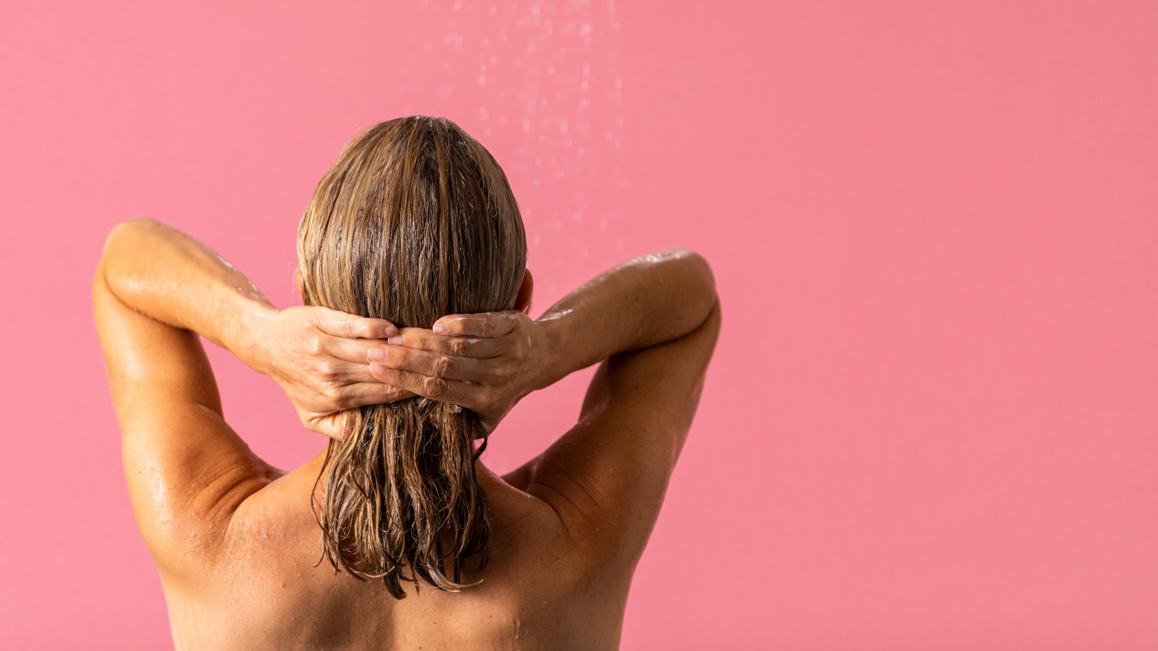 Model stands in a running shower facing away running their hands down the length of their hair on a salmon pink background.