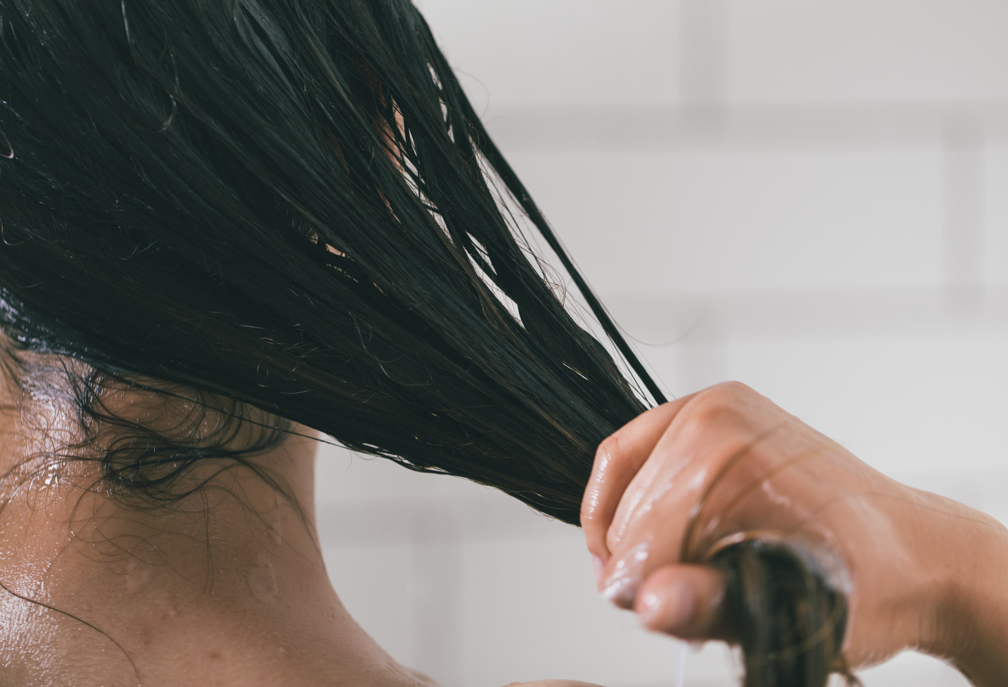 A person with long, dark, hair pulls their hair back and away from their body in a ponytail, to coat its lengths in conditioner.