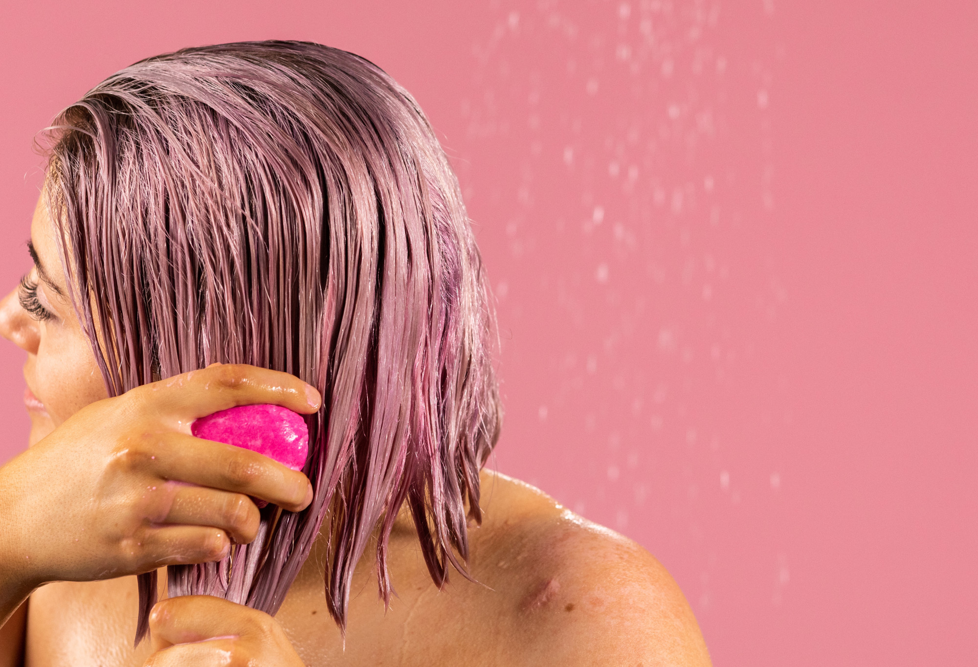 A person with bobbed pink hair works American Cream, a pink, oval shaped solid conditioner bar, through their hair.