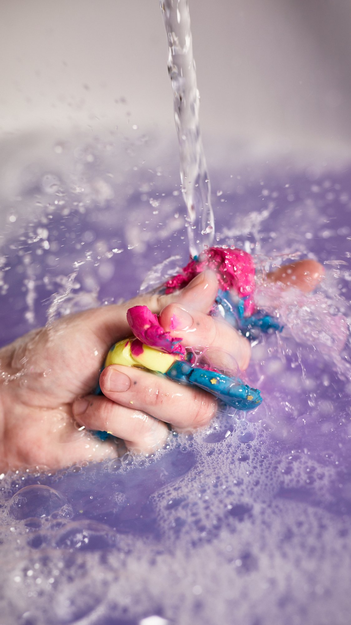 Model's hand squishes the colourful, pliable, planets under running water for extra bubbly suds. 