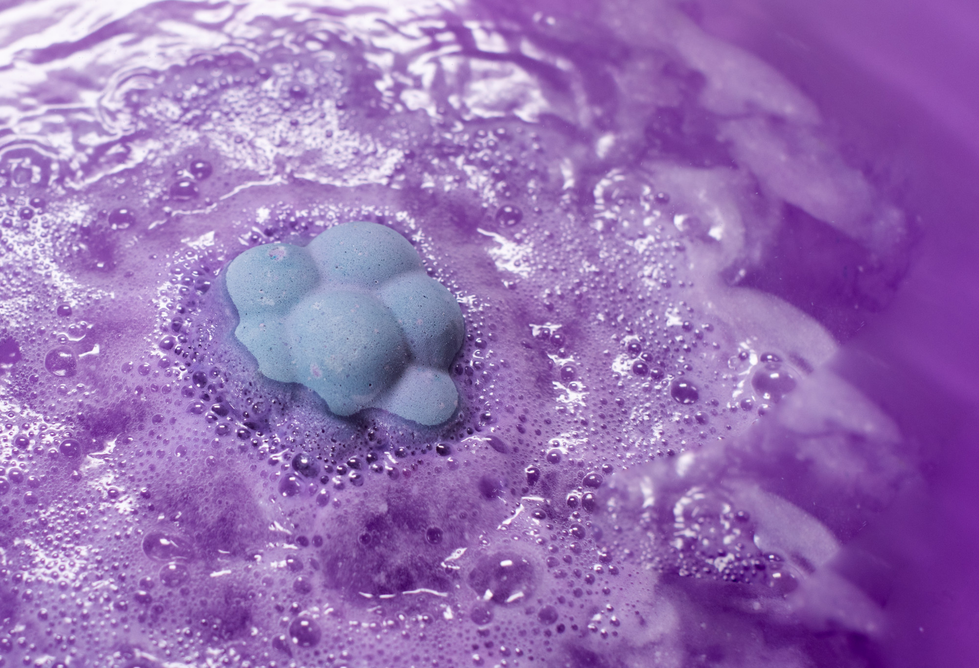 Blackberry, a baby blue, blackberry shaped bath bomb, peeking out from amethyst purple water, full of surface level bubbles.