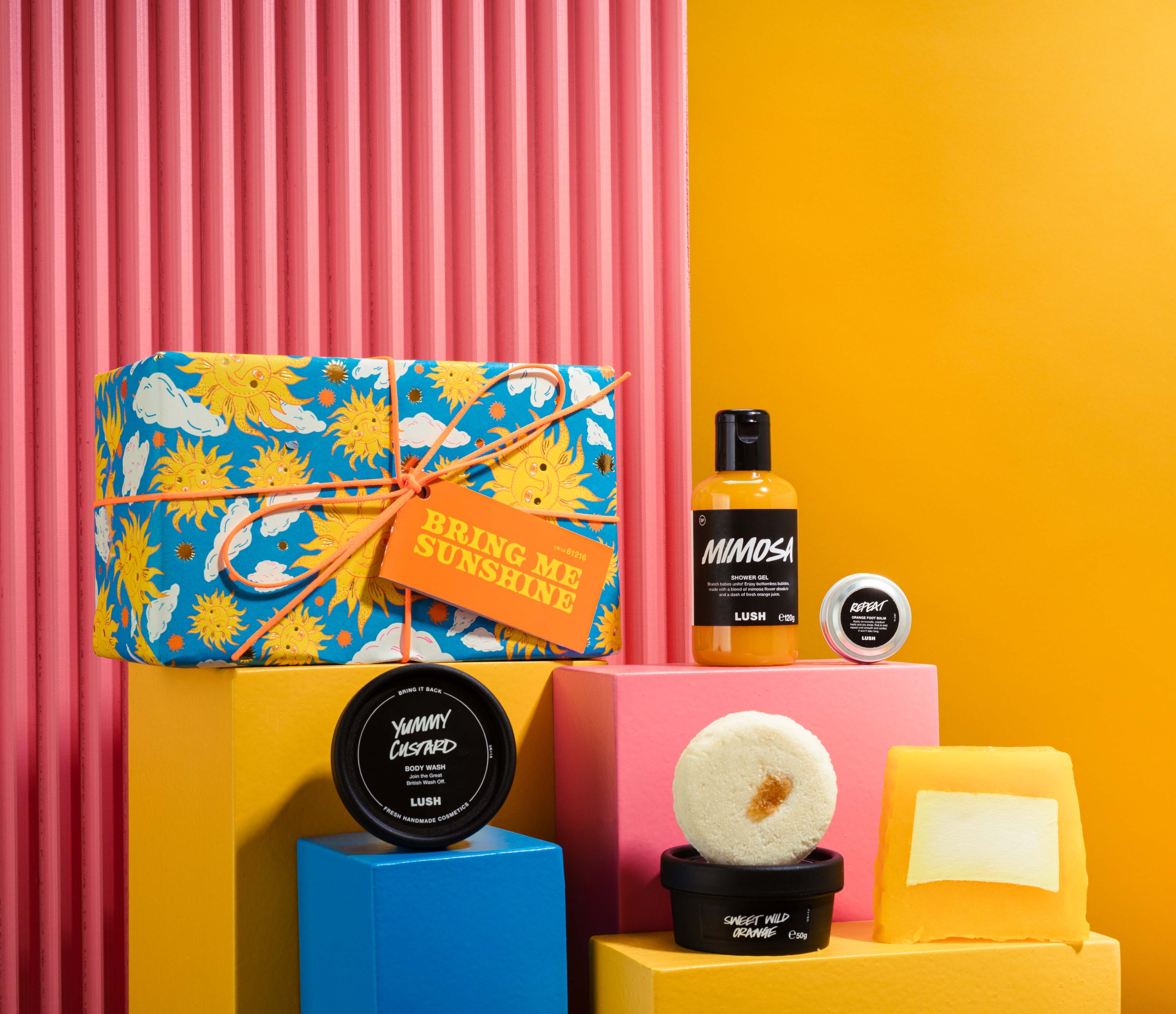Bring Me Sunshine gift, in front of a pink and yellow background, surrounded by its products on blue, yellow and pink plinths. 