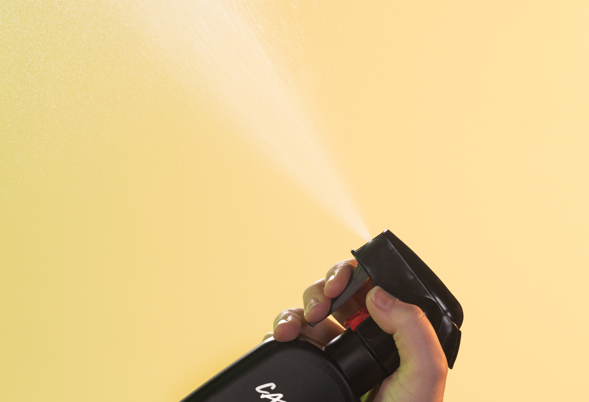 Zesty body spray is sprayed up into the air, in front of a bright, buttery yellow background.