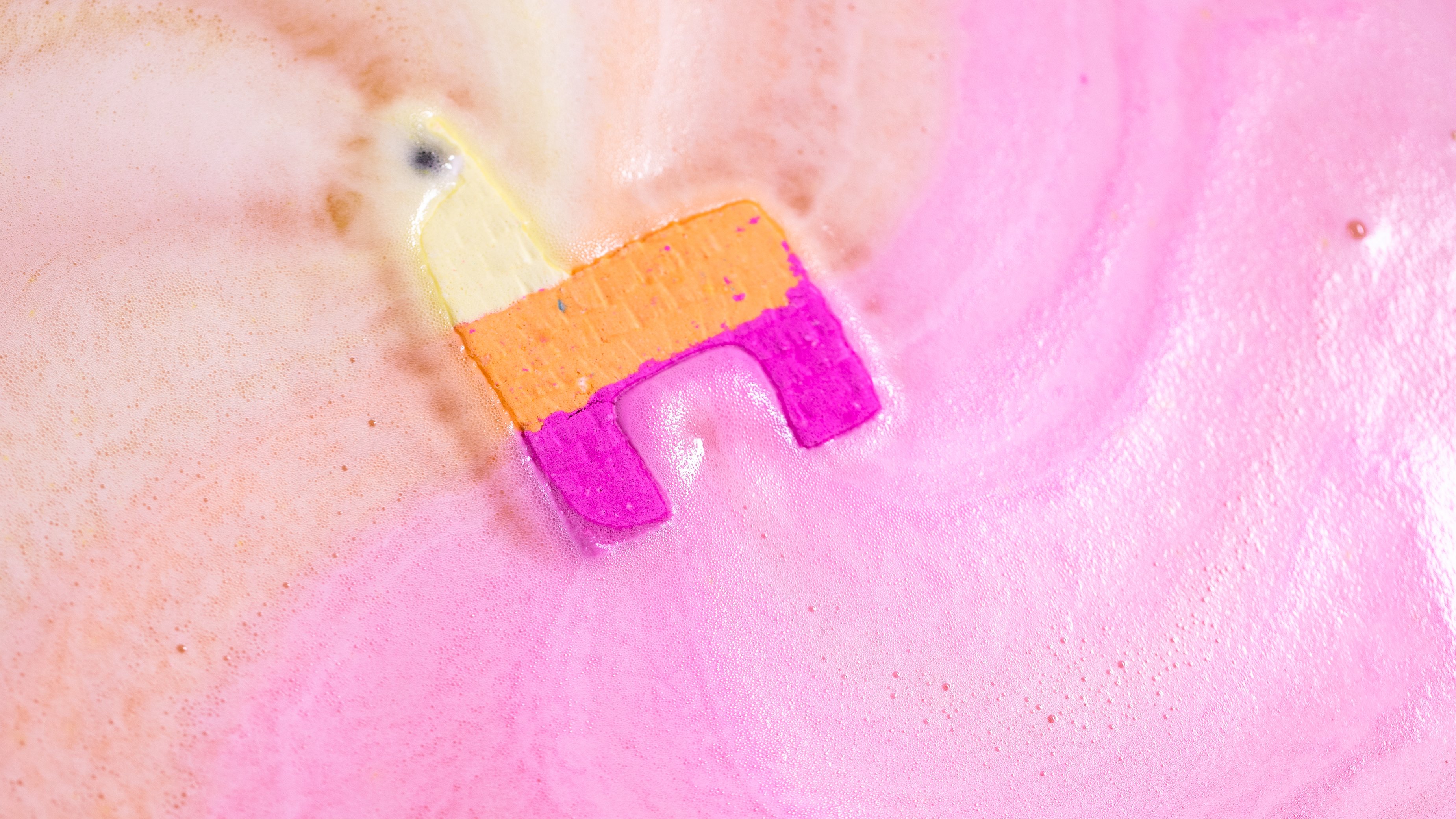 The fun, colourful, Piñata-shaped Calm A Llama bath bomb sits in the bath within foaming swirls of orange and pink.