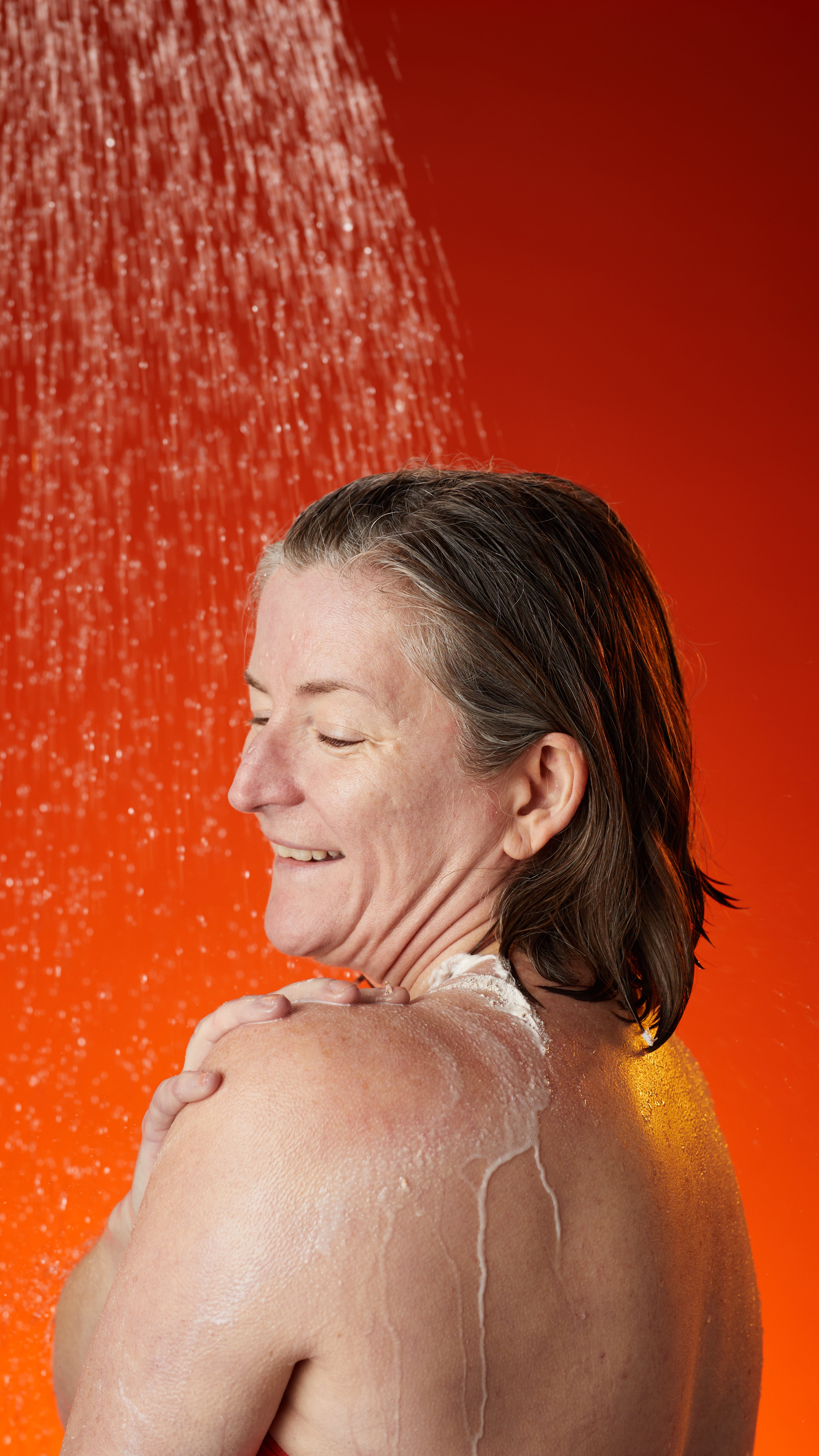 The model is showering with their back turned using the product in front of a dark to light orange gradient background.
